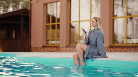 pretty-woman-is-sittign-on-edge-of-outdoor-thermal-pool-and-resting-surfing-internet-by-modern-smartphone
